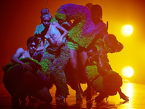A scene from the production TRAPICANA. Several performers stand close together in a dance pose, bathed in orange light.