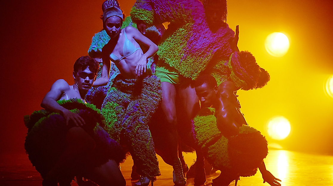 A scene from the production TRAPICANA. Several performers stand close together in a dance pose, bathed in orange light.