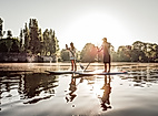 Zwei Personen beim Stand-up-Paddling im Gegenlicht auf ruhigem Wasser vor Bäumen und Abendsonne