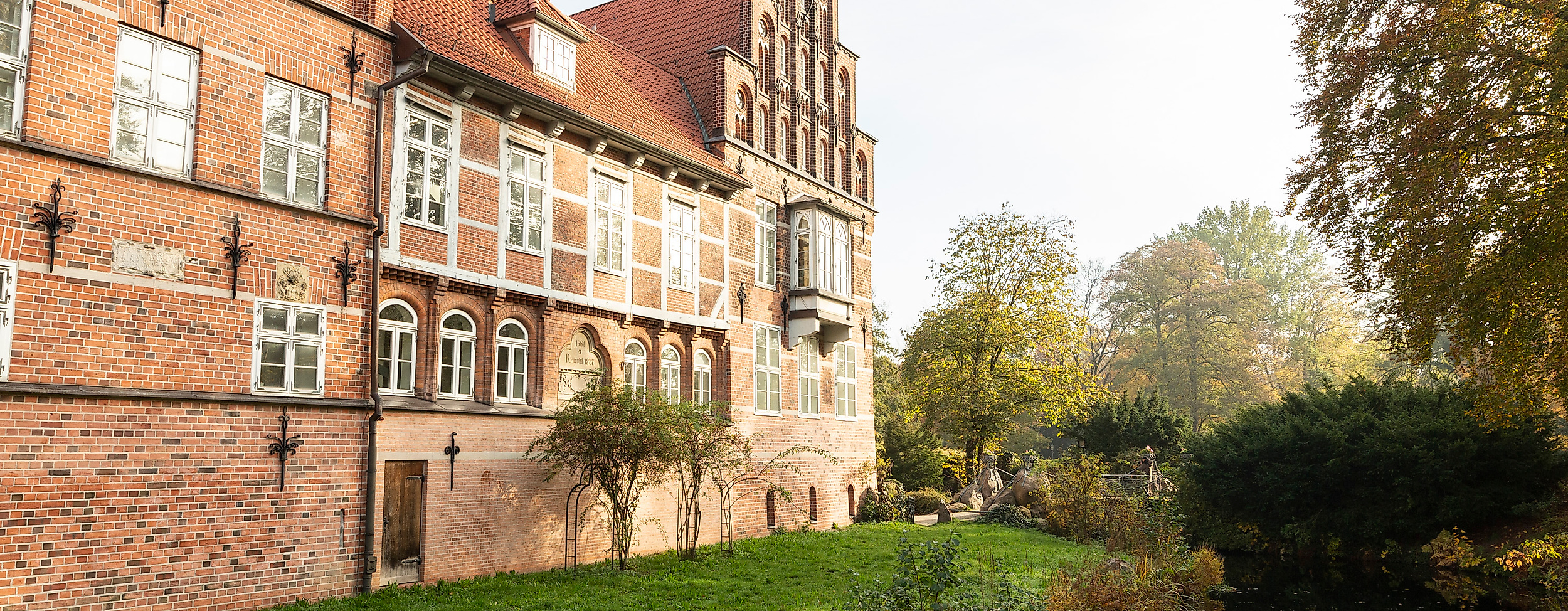 Backsteinfassade des Bergedorfer Schlosses in Hamburg mit Wassergraben und Bäumen im Sonnenlicht.