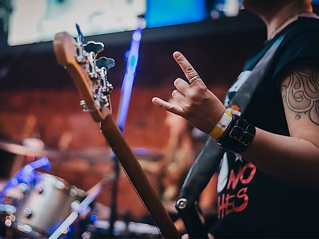 Gitarrist mit tätowiertem Arm und Rockhand-Geste auf Bühne, im Hintergrund ein Schlagzeug