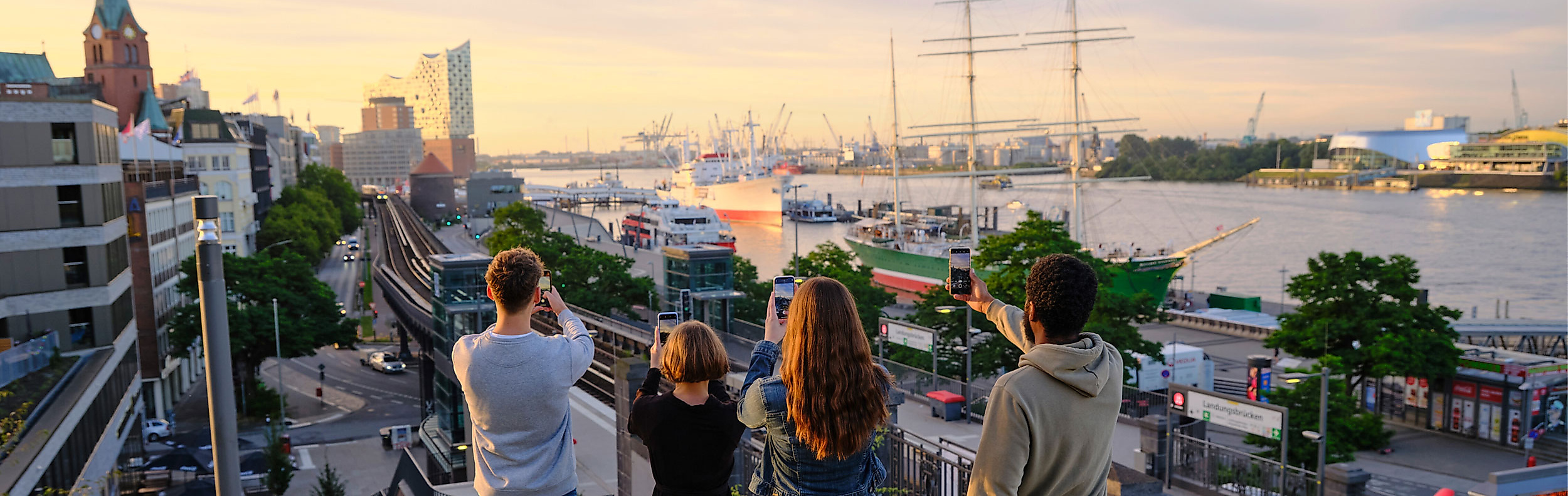 Besucher:innen fotografieren mit Handys den Hamburger Hafen und Schiffe bei Sonnenuntergang vom Landungsbrückenblick.