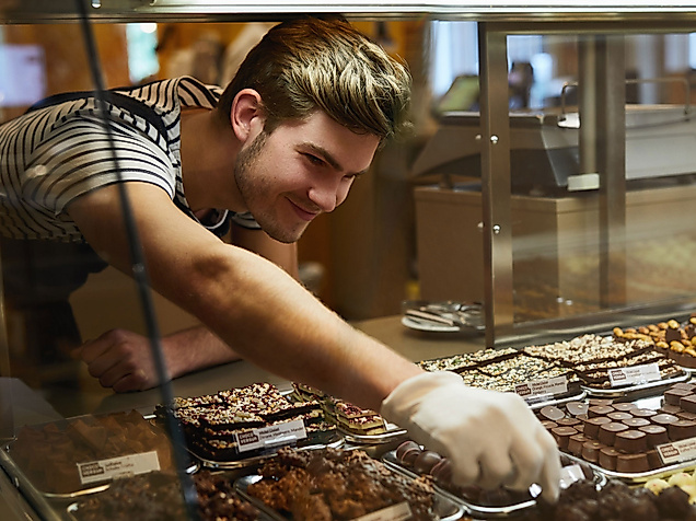 Mitarbeiter im Chocoversum greift mit Handschuh zu Pralinen hinter einer gläsernen Vitrine