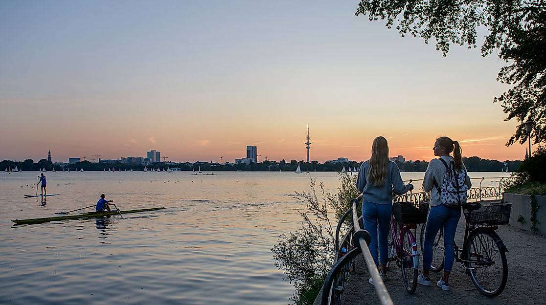 Zwei Menschen mit Fahrrädern blicken bei Sonnenuntergang auf die Alster und Hamburgs Skyline im Hintergrund