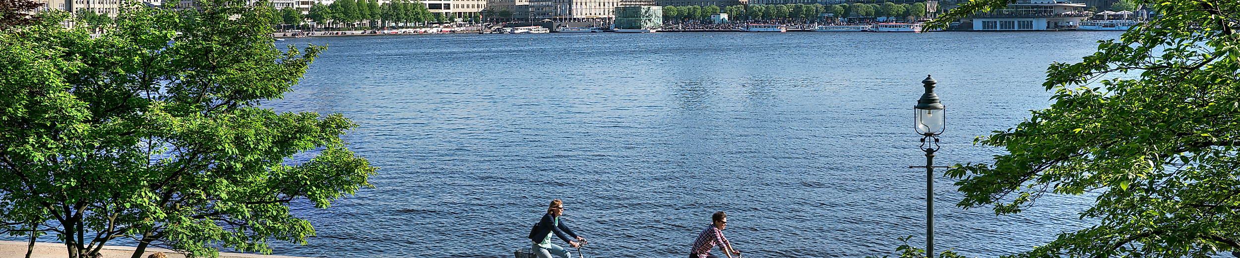 Menschen entspannen und radeln im Sommer an der Binnenalster mit Blick auf Hamburgs Skyline