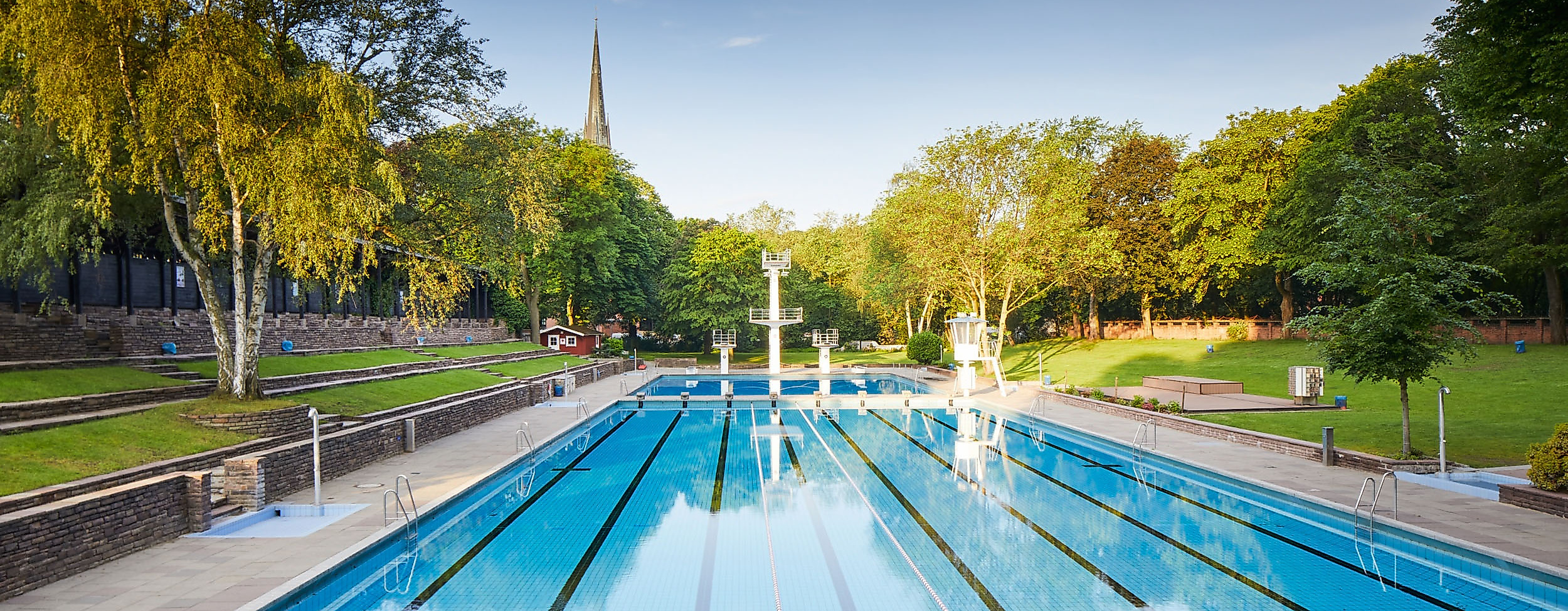 Leeres Schwimmbecken im Sommerfreibad Kaifu, umgeben von Liegewiese und Bäumen bei klarem Himmel