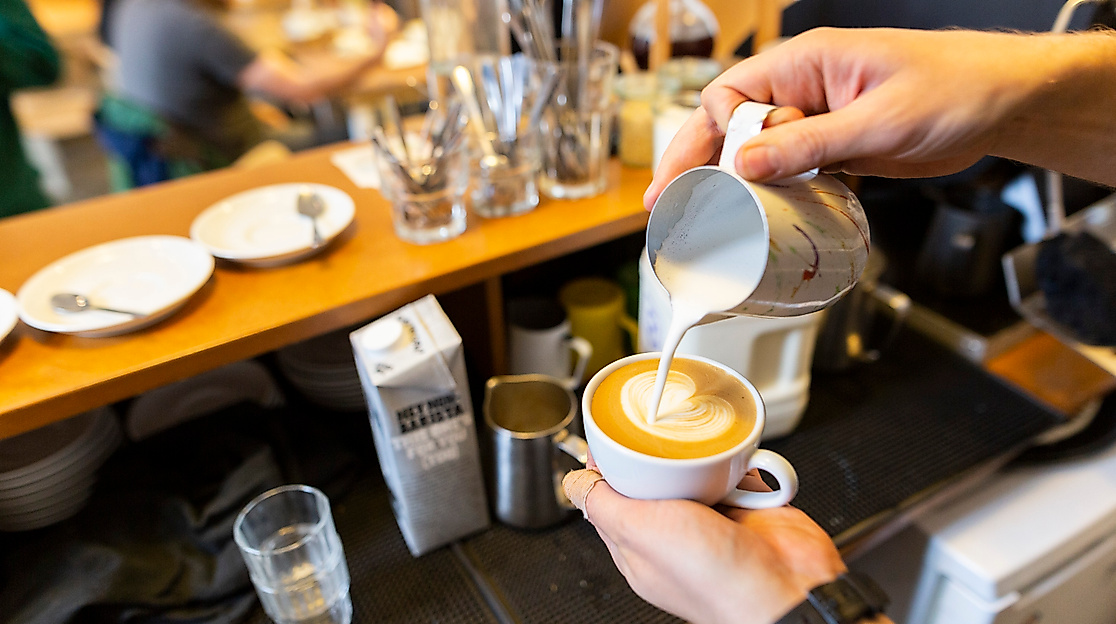 Barista schenkt Milchschaum in Cappuccino an einer Hamburger Kaffeebar ein, im Hintergrund Cafébetrieb