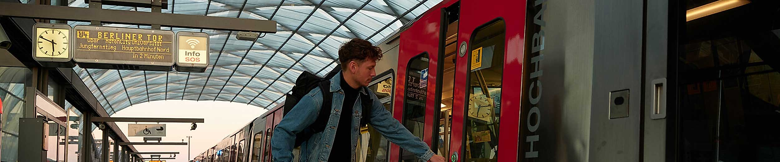 Ein junger Mann mit Rucksack steigt an einem überdachten Bahnsteig in eine rote S-Bahn ein. Auf der Anzeigetafel steht „U3 Berliner Tor“. Das Dach aus Glas lässt Tageslicht herein, im Hintergrund sind weitere Gleise und Züge zu sehen. Die Szene steht sinnbildlich für die freie und bequeme Nutzung des ÖPNV mit der Hamburg CARD