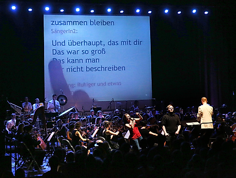 An orchestra plays on a stage, in the background is a large screen on which a song text is projected. A young woman stands in front of the orchestra, happily singing into a microphone.