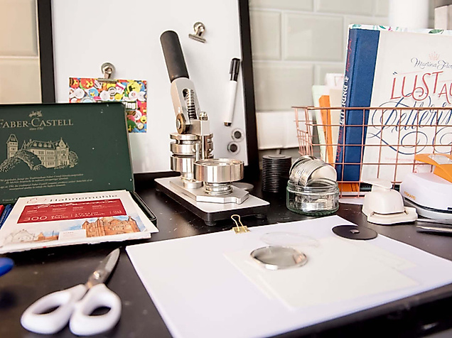 Lettering-Arbeitsplatz mit Papier, Stempelpresse, Farben, Schere und Kreativmaterialien auf schwarzem Tisch.