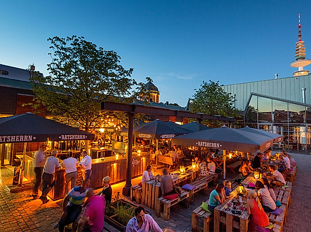 Belebter Außenbereich mit Bars und Gästen am Abend, im Hintergrund der Hamburger Fernsehturm
