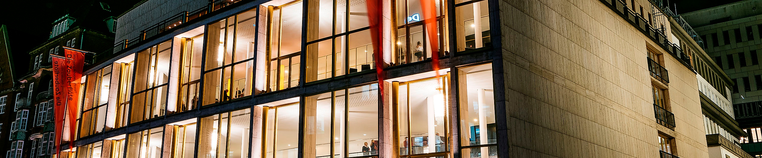 Beleuchtete Außenfassade der Hamburgischen Staatsoper bei Nacht mit Lichtspuren vorbeifahrender Autos.