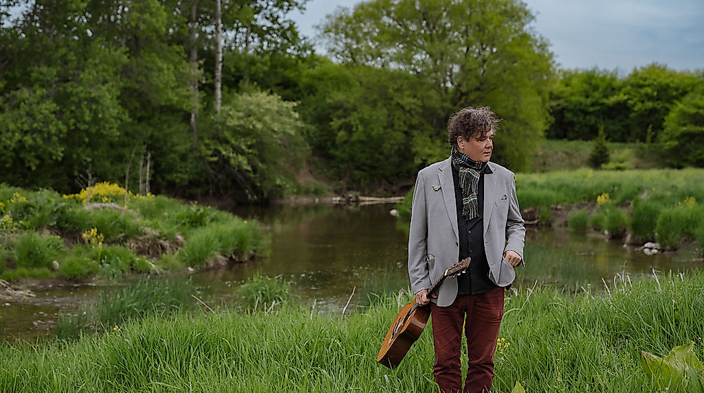 Ron Sexsmith stands in a green landscape holding his guitar.