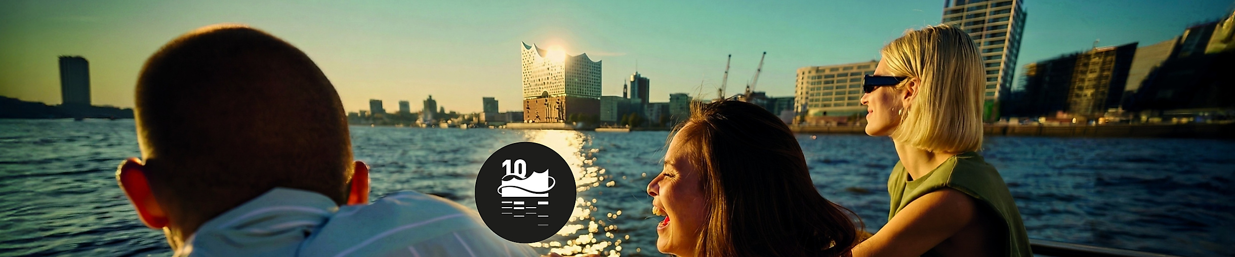 Drei Personen fahran auf einer Barkasse auf der Elbe im Hinterund ist die Elbphilharmonie zu sehen