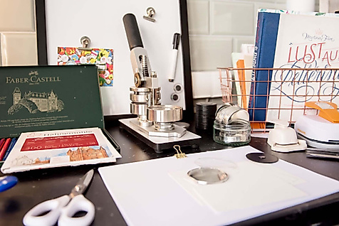 Lettering-Arbeitsplatz mit Papier, Stempelpresse, Farben, Schere und Kreativmaterialien auf schwarzem Tisch.