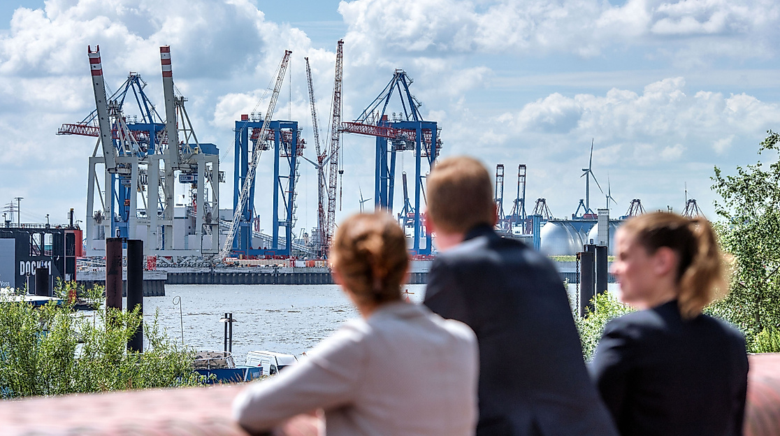 Drei Personen blicken bei Sonnenschein auf Containerkräne im Hamburger Hafen