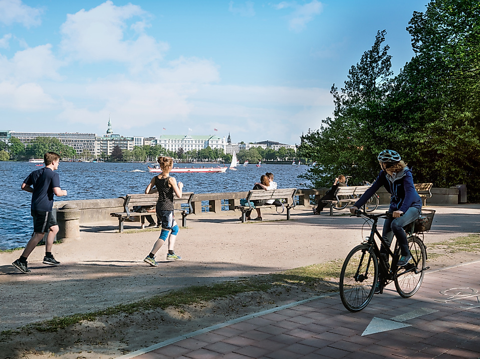 Radfahrer und Jogger am sonnigen Uferweg der Alster mit Blick auf Wasser und Stadtpanorama
