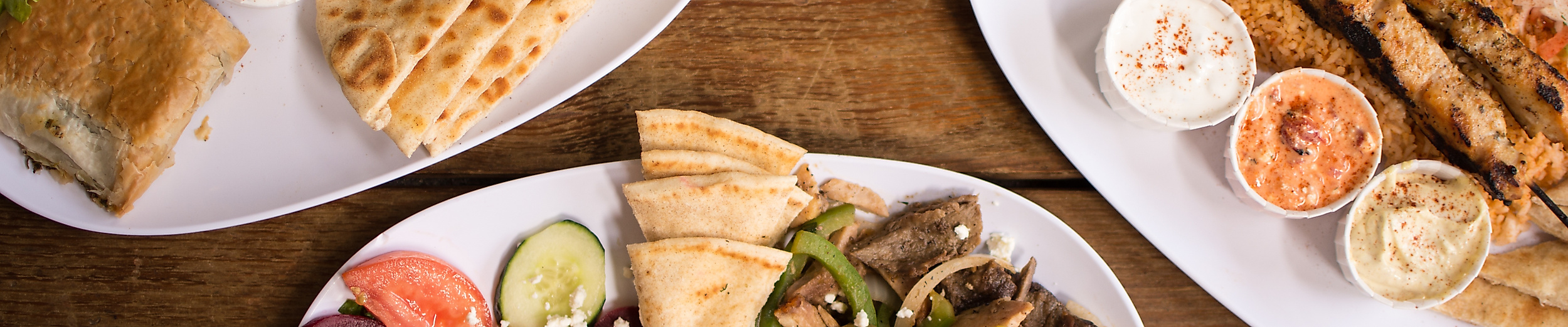 Griechische Spezialitäten wie Gyros, Souvlaki, Tzatziki und Fladenbrot auf drei Tellern serviert.