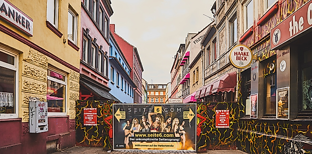 Blick in die Herbertstraße mit Sichtschutzwand, flankiert von bunt beleuchteten Fassaden und Schildern auf der Reeperbahn