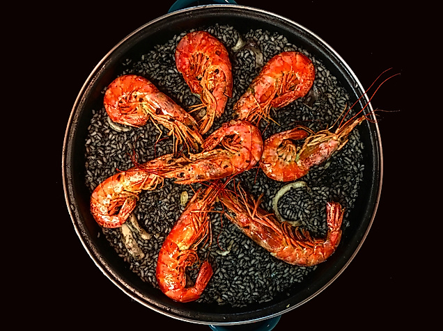 Pfanne mit schwarzer Paella und acht großen Garnelen auf dunklem Hintergrund stilvoll angerichtet.