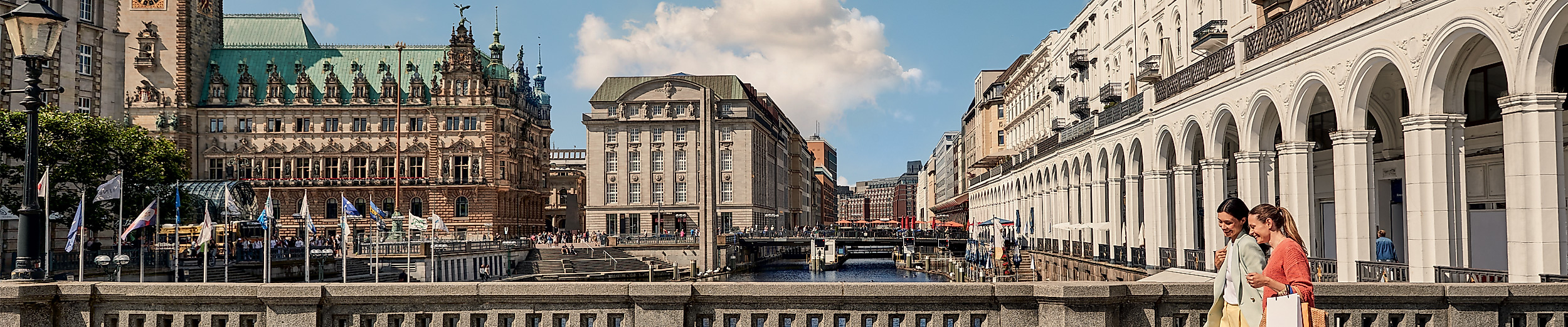 Blick auf Hamburgs Alsterarkaden mit Einkaufsflair und Rathaus im Hintergrund bei Sonnenschein.