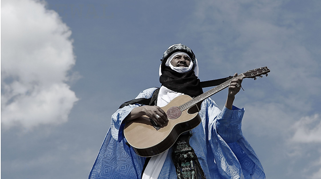 Alhousseini Anivolla is standing on a rooftop with a guitar. The sky can be seen in the background.