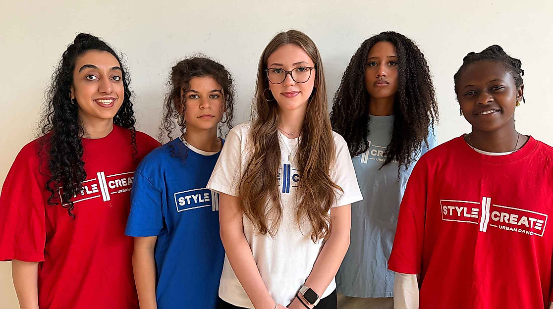 Five people with different hair lengths and textures are wearing colorful T-shirts with the words “STYLE CREATE” printed on them and are standing in front of a light-colored wall.