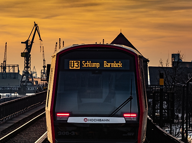 U-Bahn der Linie U3 Richtung Barmbek bei Sonnenuntergang, mit Hafenkränen im Hintergrund.