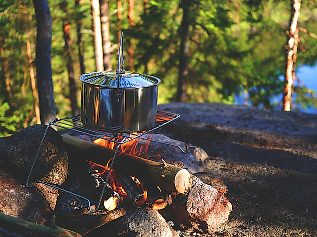 Kochtopf über offenem Feuer auf Holzunterlage im Wald nahe einem See beim Camping