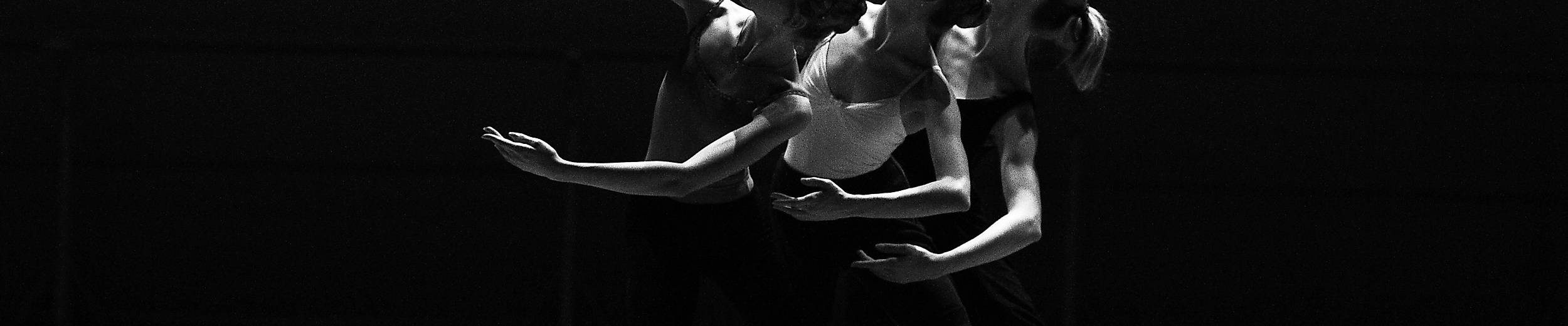 Drei Tänzer:innen in synchroner Bewegung auf dunkler Bühne, festgehalten in dynamischer Schwarzweißaufnahme