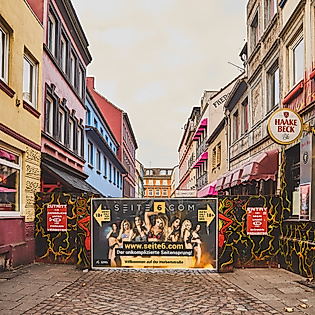 Blick in die Herbertstraße mit Sichtschutzwand, flankiert von bunt beleuchteten Fassaden und Schildern auf der Reeperbahn