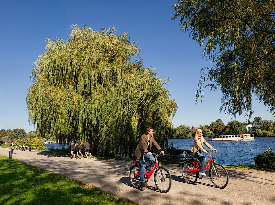 Zwei Personen radeln mit Stadträdern am Ufer der Alster entlang unter blauem Himmel.