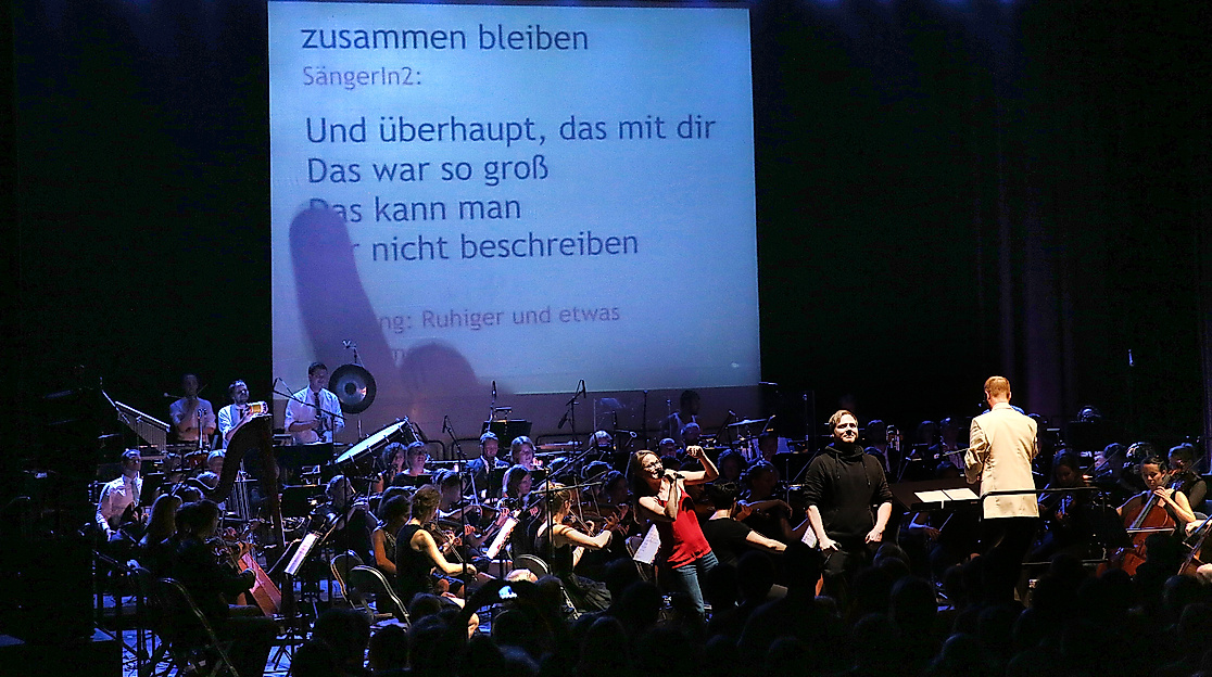 An orchestra plays on a stage, in the background is a large screen on which a song text is projected. A young woman stands in front of the orchestra, happily singing into a microphone.