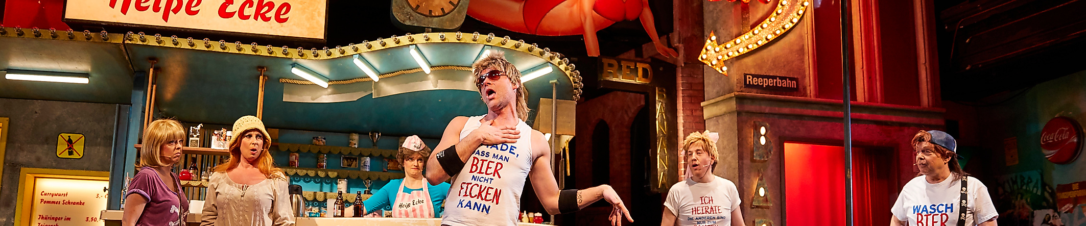 Szenenbild Heiße Ecke: 5 Darsteller:innen in schrillen Kostümen vor einem Imbissstand und leuchtenden Reklametafeln