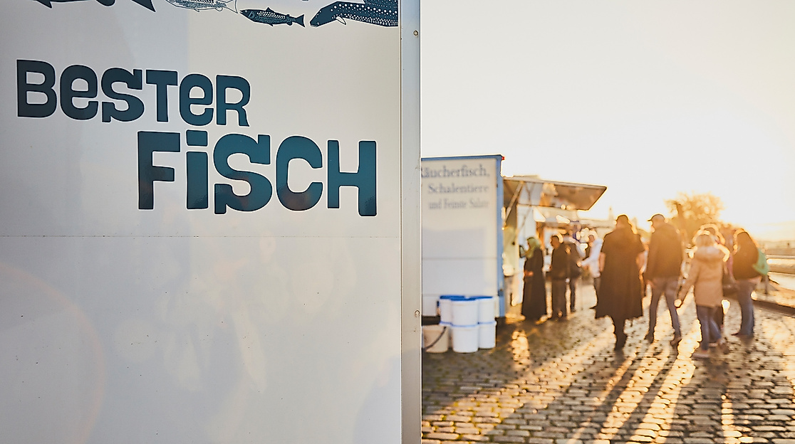 Schriftzug „Bester Fisch“ am Stand des Hamburger Fischmarkts, Menschen im Morgenlicht auf Kopfsteinpflaster