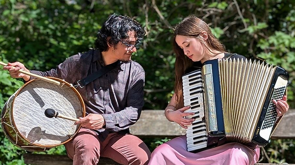 Víctor Gutiérrez and Isidora Caroca are sitting on a bench. While Víctor is holding a percussion instrument, Isidora is holding an accordion.