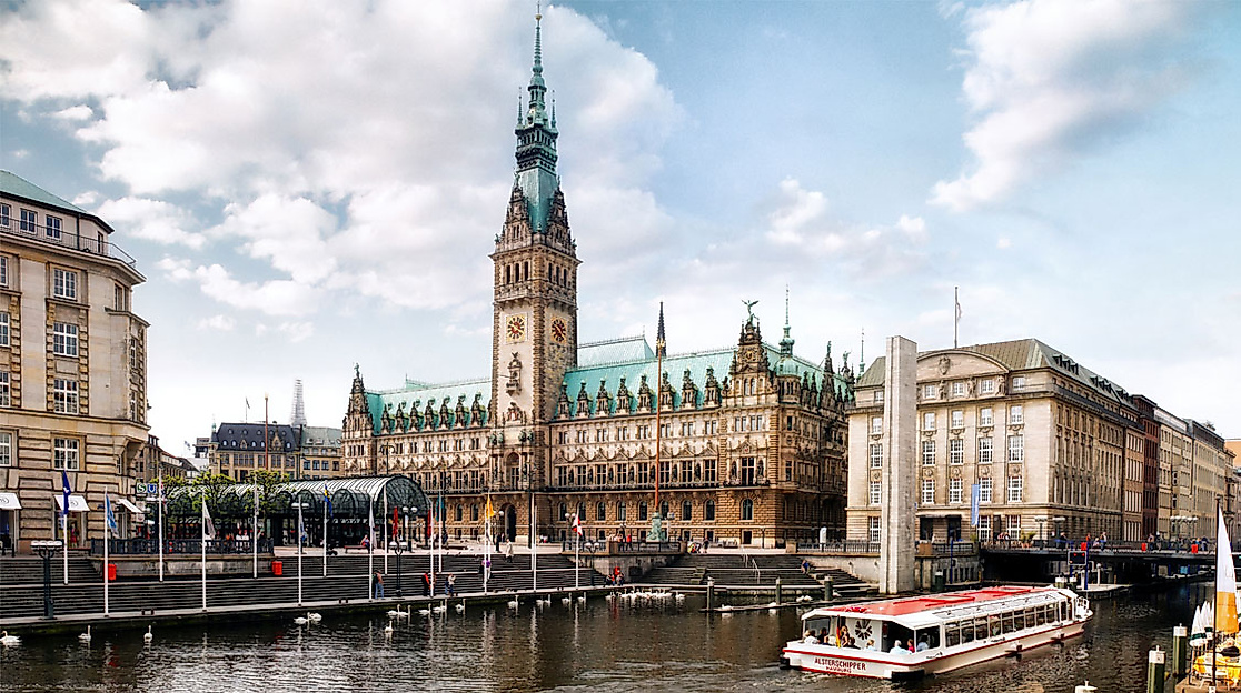 Blick auf das Hamburger Rathaus am Alsterfleet mit Ausflugsbooten, historischen Fassaden und blauem Himmel.