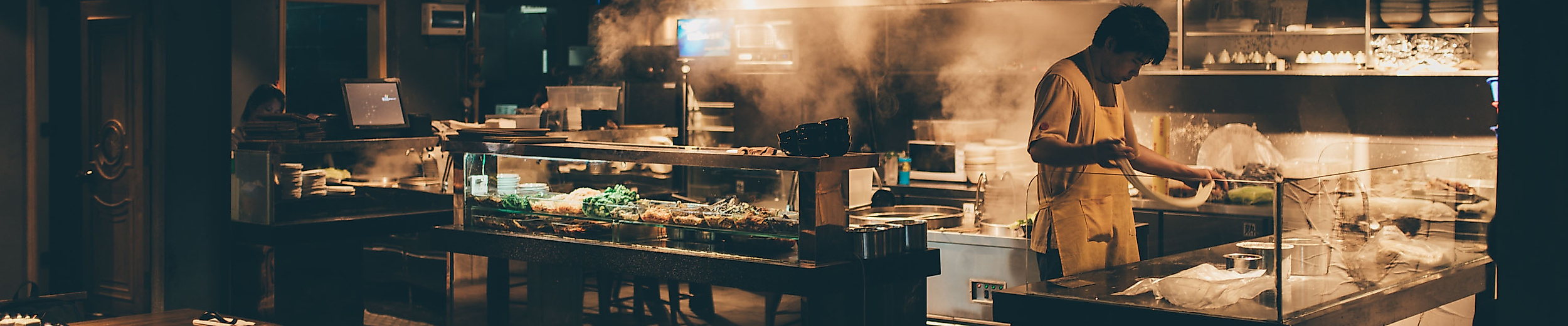 Offene Restaurantküche mit Köchen bei der Zubereitung, im Vordergrund gedeckte Tische in warmer Beleuchtung