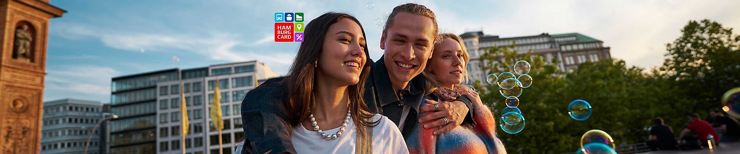 Drei junge Menschen genießen gemeinsam einen sonnigen Moment auf dem Platz vor der Hamburger Kunsthalle. Sie lachen, posieren eng umarmt und sind umgeben von schwebenden Seifenblasen. Im Hintergrund moderne und historische Gebäude, darüber ein weiter Himmel. Die Szene versprüht Lebensfreude, Leichtigkeit und Vorfreude auf gemeinsame Erlebnisse in Hamburg.