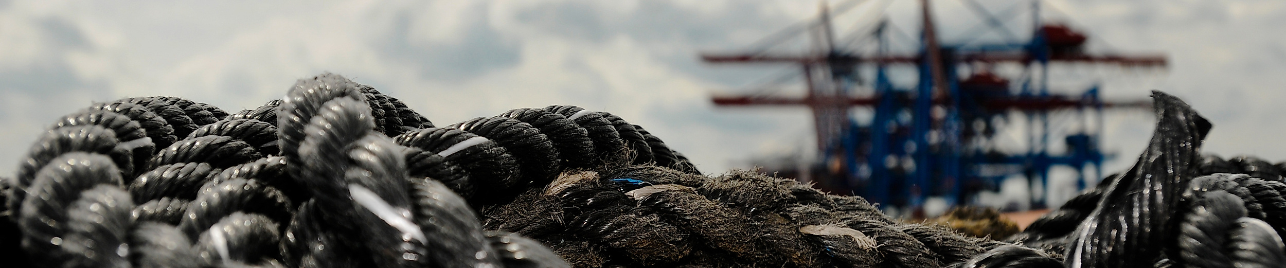 Vor einem verschwommenen Containerschiff im Hintergrund des Bildes, das vor dem bewölkten Himmel zu sehen ist, liegt ein dickes, graues Tau auf dem Boden