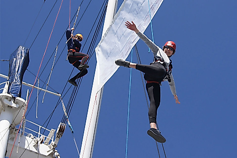 Zwei gesicherte Personen klettern im Hochseilgarten auf der Cap San Diego vor blauem Himmel
