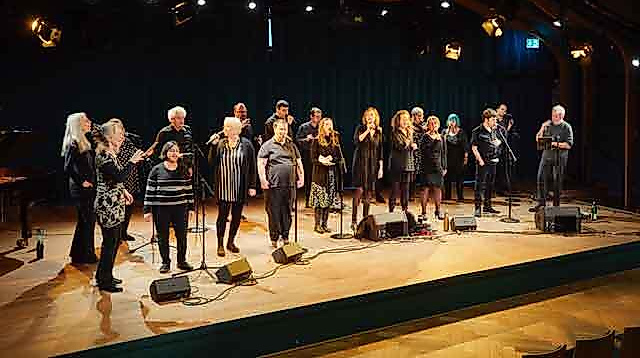 A group of people is standing on a stage, singing together into microphones.