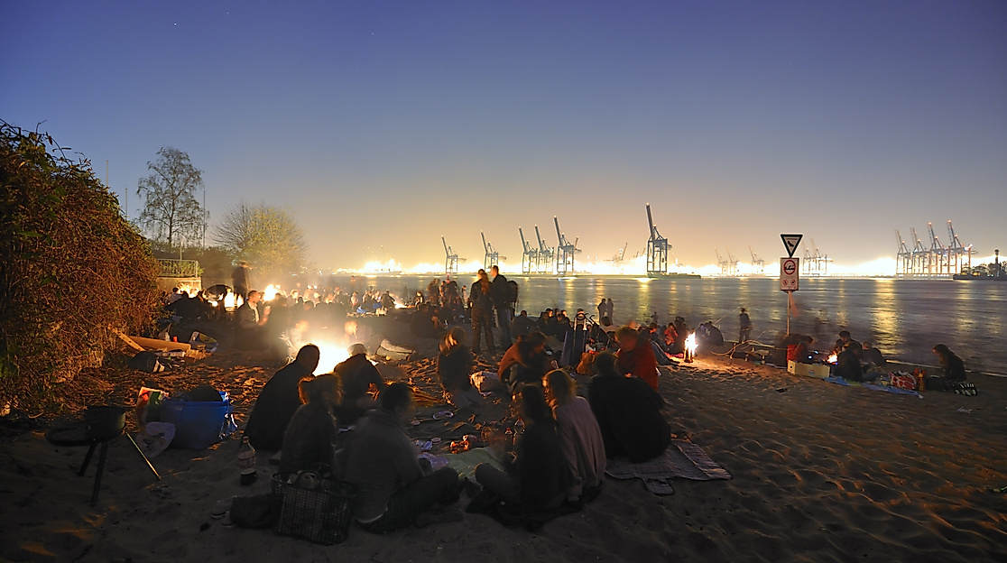 Menschen feiern Osterfeuer am Elbstrand bei Nacht mit Blick auf den beleuchteten Hamburger Hafen