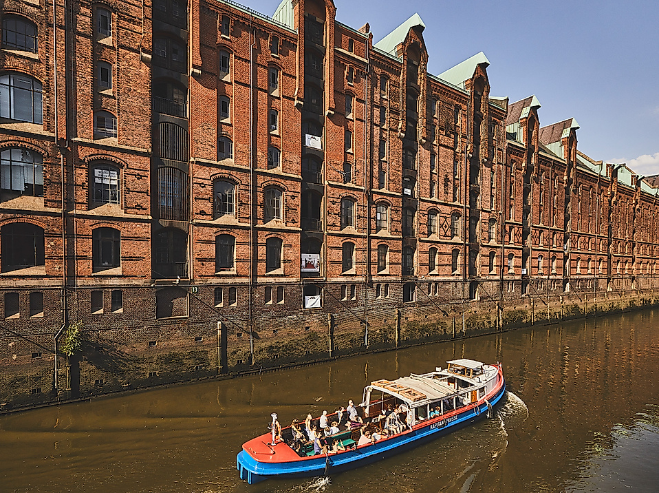 Barkasse fährt durch einen Fleet der Speicherstadt entlang roter Backsteinfassaden bei Sonnenschein