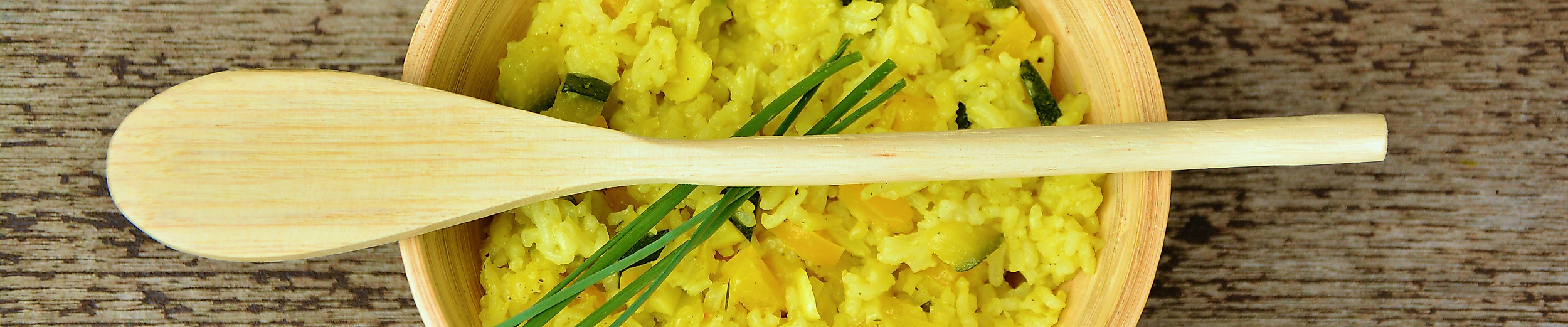 Schüssel mit gelbem Curryreis, garniert mit Schnittlauch, dazu Holzlöffel auf rustikalem Holztisch