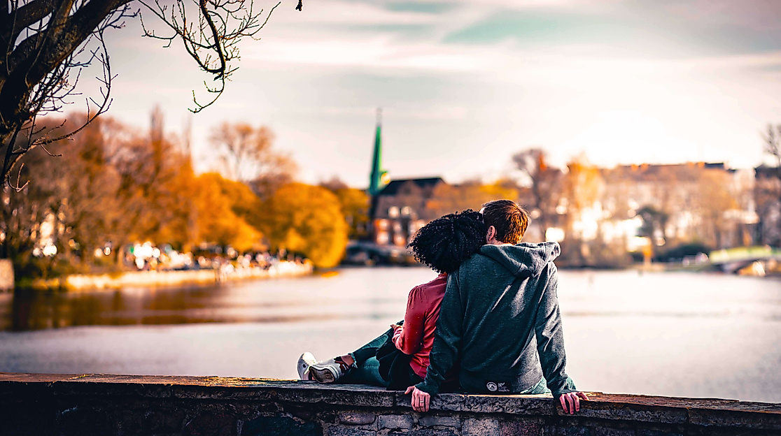 Paar sitzt eng umschlungen an der Alster mit Blick auf herbstlich gefärbte Uferlandschaft