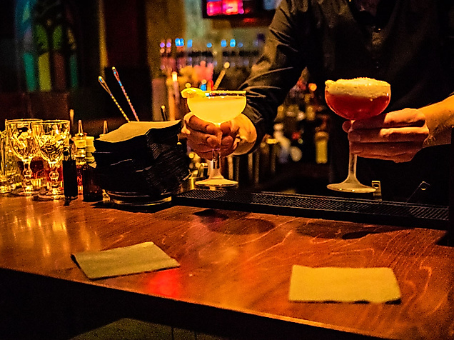 Barkeeper serviert farbenfrohe Cocktails an dunkler Theke in Hamburger Bar bei stimmungsvollem Licht.