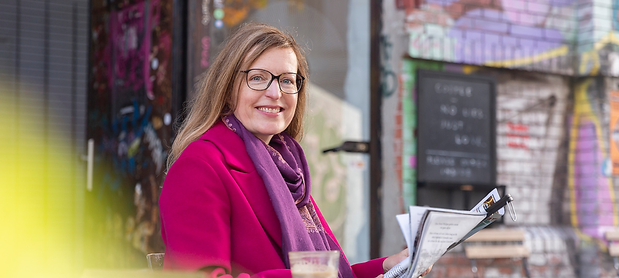 Lächelnde Frau mit Zeitschrift und Getränk im Straßencafé vor bunt besprühter Wand im Hamburger Schanzenviertel.