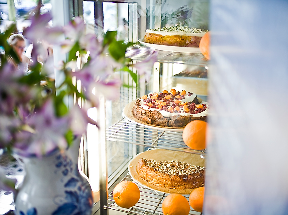 Kuchenvitrine mit frischen Torten und Orangen im Café Johanna, daneben Frühlingsblumen in einer Vase