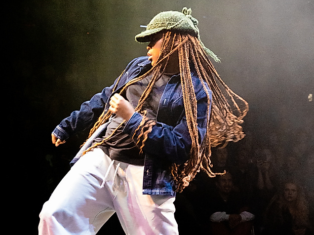 A person wearing a knitted hat, long braids, and loose clothing stands on a stage and moves their hips in one direction with bent knees. They pull themselves up in the same direction with the opposite arm.
